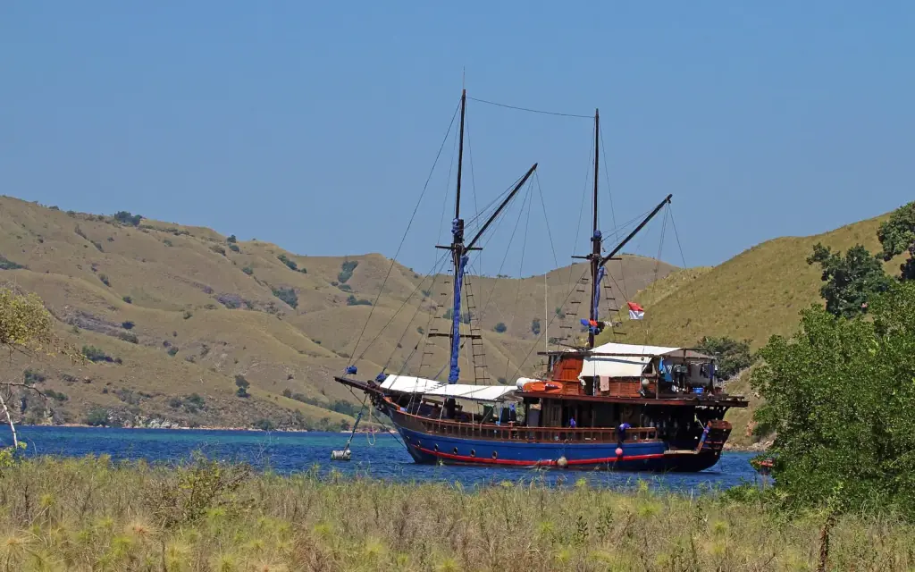 A two-masted wooden charter boat moored alone in a quiet bay at Lawa Darat, seen across a grassy hillside.
