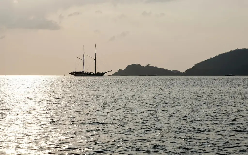 A two-masted schooner silhouetted on silver water between islands off Labuan Bajo.