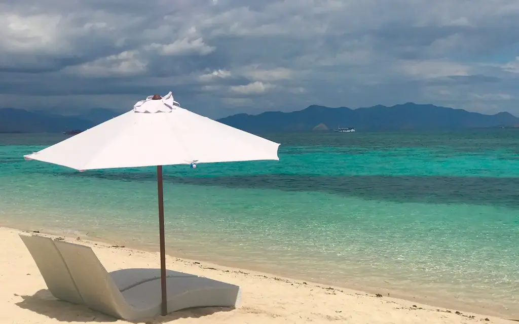 A white parasol and sun lounger on the sand at Kanawa Island, facing the shallow turquoise reef.