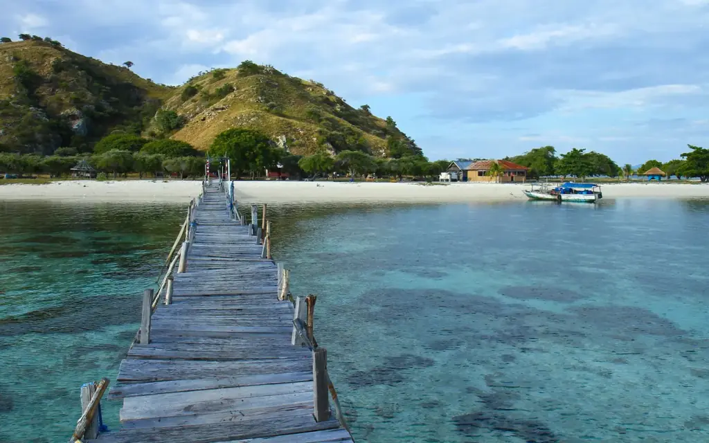 The wooden jetty that used to run out over the reef at Kanawa Island, photographed before resort management dismantled it in August 2024; the island beach and hill behind, Komodo National Park