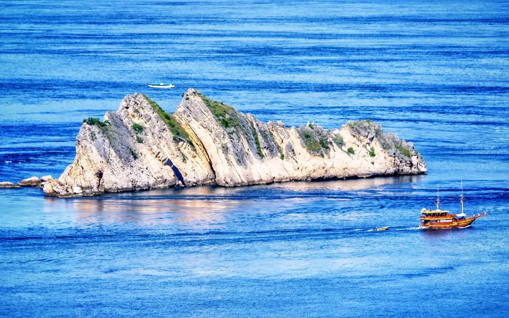 A steep white limestone islet rising from deep blue water near Labuan Bajo, with a wooden boat passing alongside.