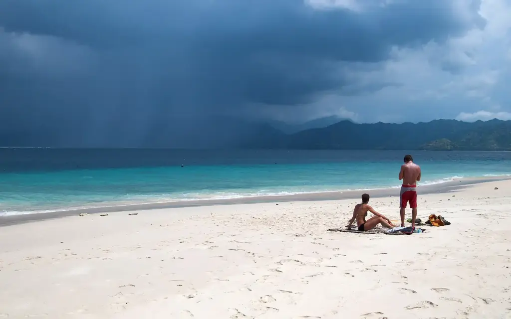 Two people sitting on a white-sand beach as a rain squall crosses the channel behind them, Gili Meno.