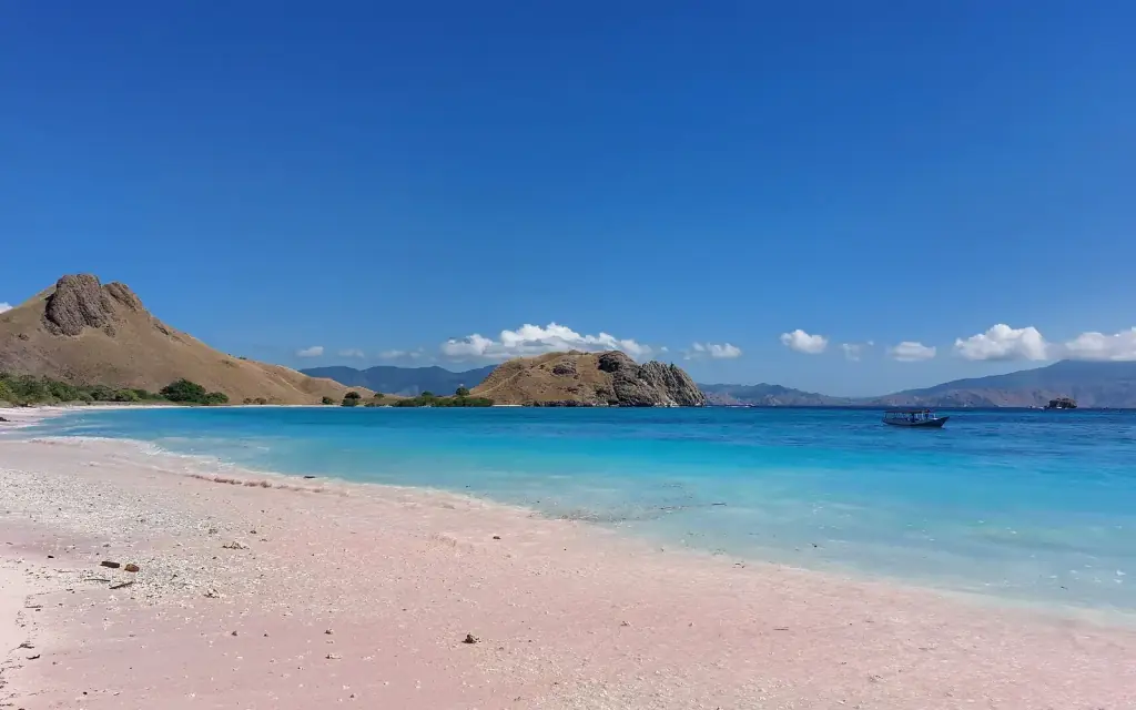 Pink-tinted sand meeting pale turquoise shallows at Pink Beach, with dry hills across the bay.