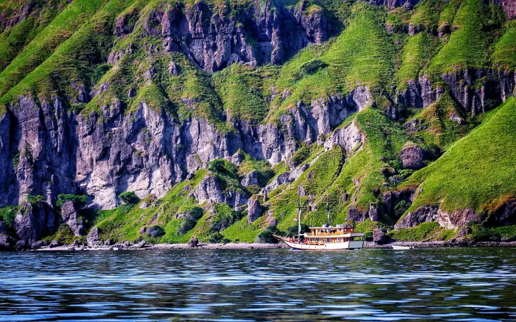 A wooden phinisi passing beneath steep green cliffs in the waters around Labuan Bajo.