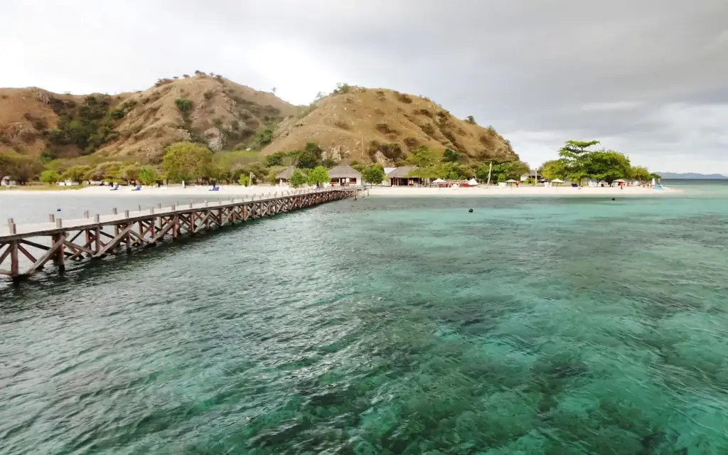 Dermaga kayu Pulau Kanawa yang menjorok ke laut dangkal, dengan pantai dan bukit pulau di belakangnya, Flores, Nusa Tenggara Timur.