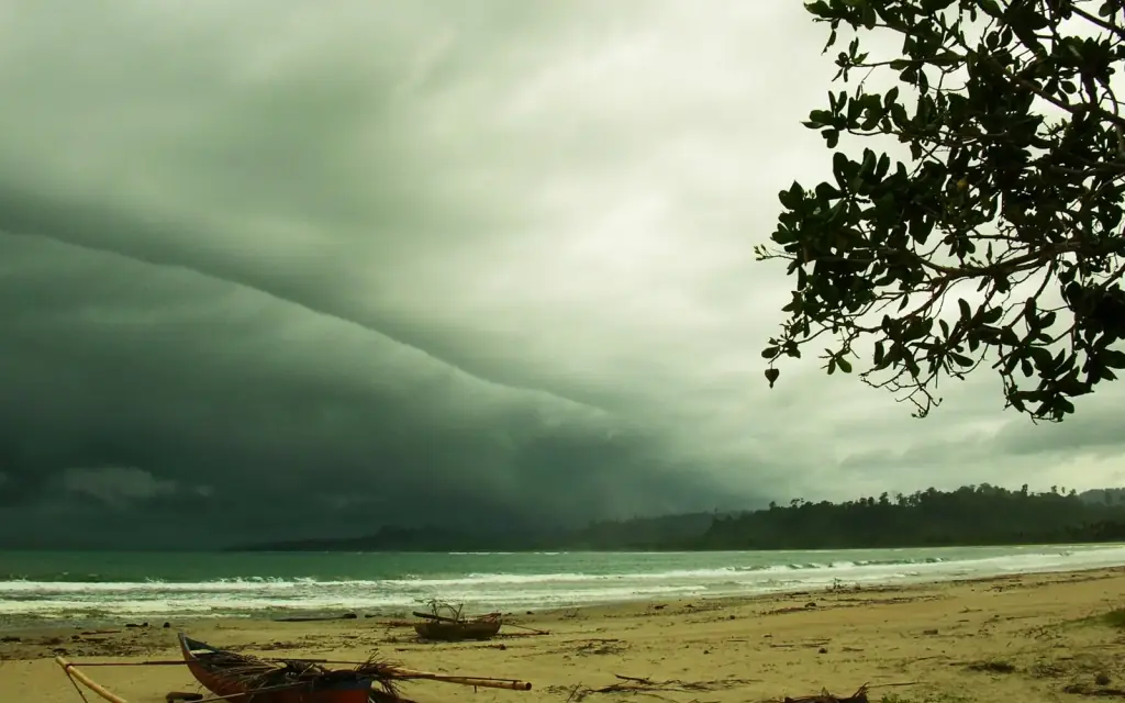 A heavy storm front rolling in over the sea above a beach with outrigger canoes drawn up on the sand.